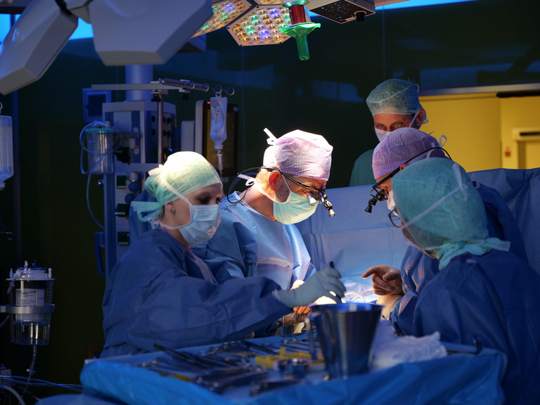 Prof. Dr. Hanke steht mit dem OP-Team im tiefblau erleuchteten Operationssaal und blickt mithilfe einer LED-Stirnlampe auf das Operationsfeld während einer Herz-Operation im Albertinen Herz- und Gefäßzentrum in Hamburg Prof. Dr. Hanke steht mit dem OP-Team im tiefblau erleuchteten Operationssaal und blickt mithilfe einer LED-Stirnlampe auf das Operationsfeld während einer Herz-Operation im Albertinen Herz- und Gefäßzentrum in Hamburg